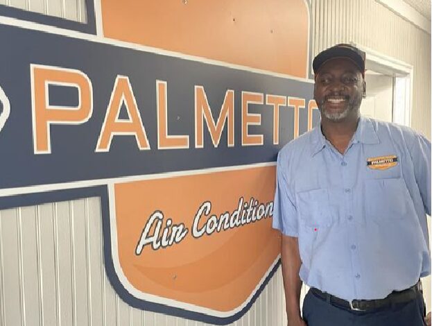 A man wearing a blue uniform and cap is smiling in front of a sign that reads "Palmetto Air Conditioning." The sign has an orange and blue color scheme.