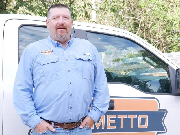 A man with short hair and goatee stands in front of a white truck wearing a light blue button-up shirt and jeans. The truck door has a logo partially visible with the word "METTO" on it. Trees are in the background.